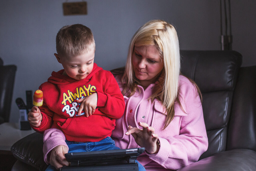 Little boy eating an ice lolly sits on his mum's knee as they watch a video on an iPad together. 