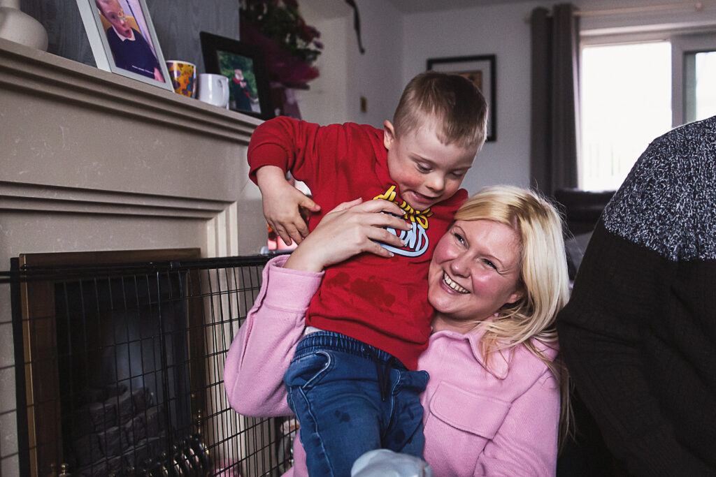 A woman wearing a pink jacket is sat on the floor holding up her son who is in a red hoodie.