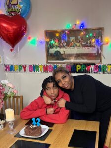 Shantai and Derion are next to a dining table, in a room decorated with birthday banners and balloons. Derion is wearing red, looking at the camera. Shantai has her arms around Derion.