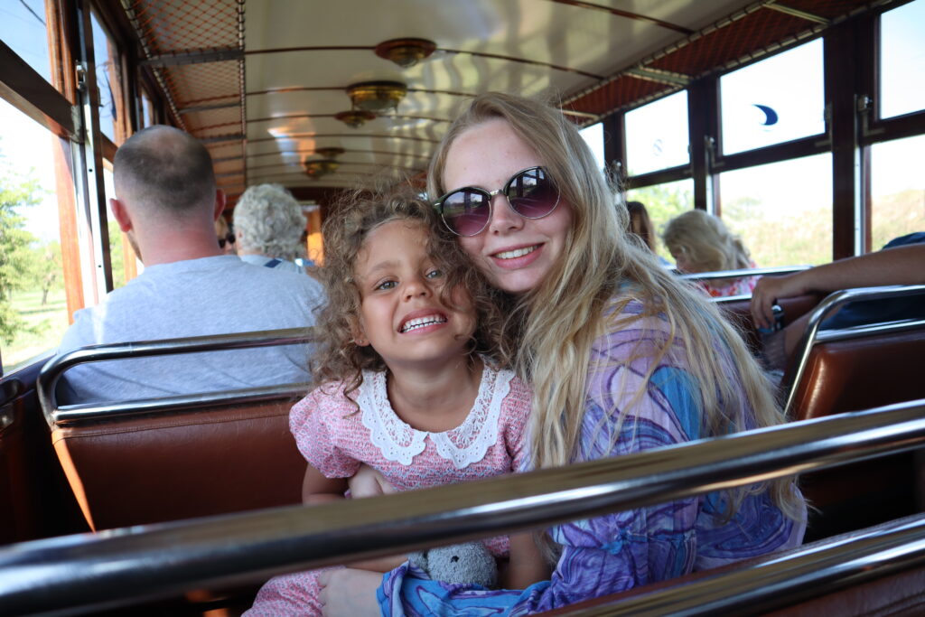 Lauryn and Amelia are smiling at the camera. They are sitting on a bus, both turning to face the camera. It is sunny outside and they're wearing dresss. Amelia has dark hair, Lauryn has blonde hair and her arm around Amelia. Amelia is holding a teddy.