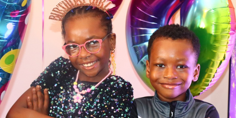 Elizabeth and Testimony are standing side by side with their arms crossed and smiling. Elizabeth is wearing a 'birthday girl' headband, and there are balloons in the background.