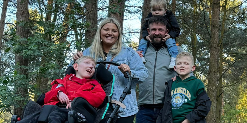 Mason, his brothers and parents are at Centre parcs walking amongst the trees on a path. They are all smiling at the camera. Mason is in a specialist pram, and Arthur is on Kris' shoulders. Archie is stood next to Kris, who has his hand on his shoulder. Jess is pushing Mason.