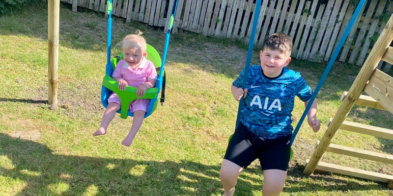 Parker is playing on the swing set from Family Fund with his little sister Nala-Reign. They are both smiling and mid-swing.