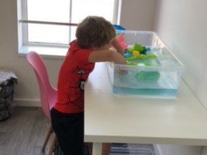 A little boy in a red t-shirt is sat at a white table with his hands in a large clear tub with water and toys in