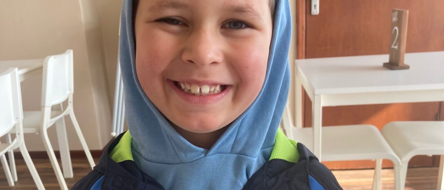Cody is sat in a cafe with a slice of rainbow cake in front of him. He is smiling widely at the camera, wearing a hood. He has dark brown hair.