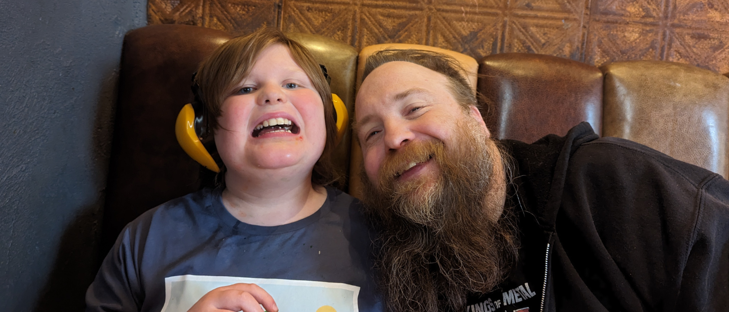 Connor is sat at a table wearing headphones and holding felt-tips, next to his dad Kris. Kris is leaning into him and they are both smiling.