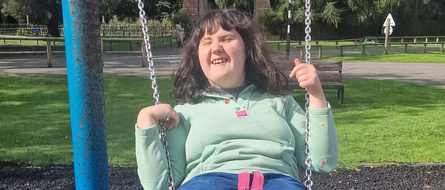 Kirsten is smiling whilst sat on a swing in a park. She is holding onto the chains and leaning back.
