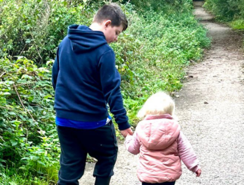 Parker is holding his little sister Nala-Reign's hand. They are walking down a woodland path with their backs to the camera.
