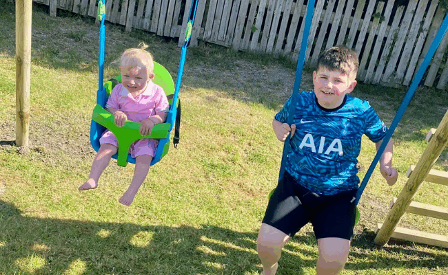 Parker is playing on the swing set from Family Fund with his little sister Nala-Reign. They are both smiling and mid-swing.