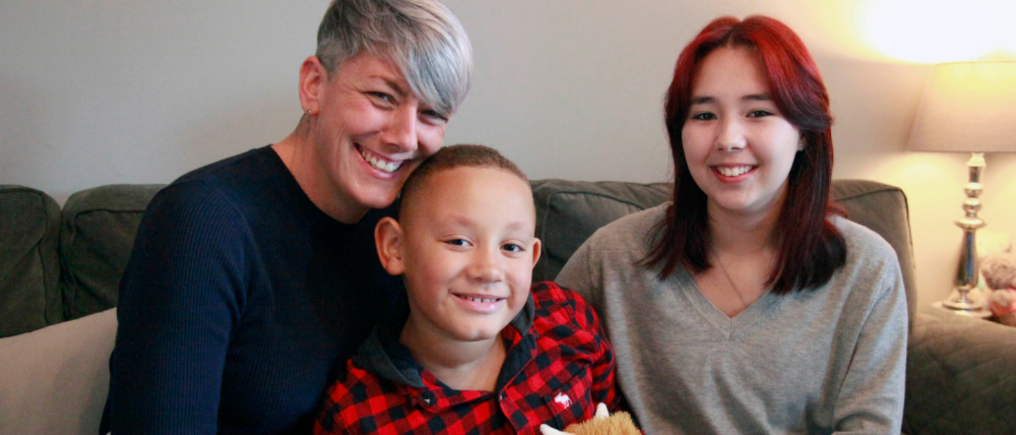 A photo of a mother sitting on the sofa next to her son and daughter. They are all smiling at the camera.