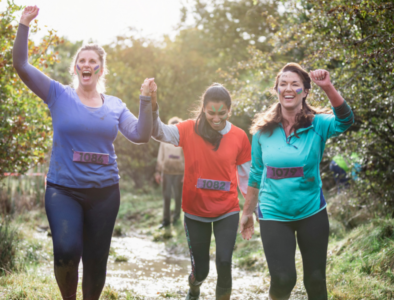 Three women are running in mud holding hands and smiling.
