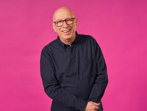 A bright pink background with a man smiling. He's wearing a dark navy shirt and square glasses.