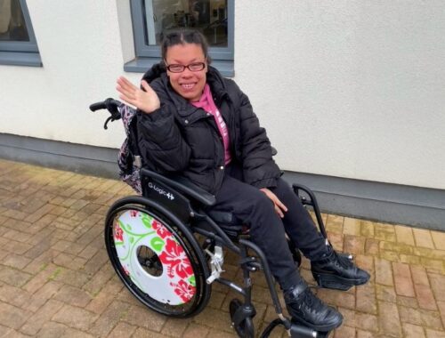 A young woman outside in a wheelchair smiling and waving