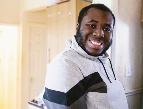 Anjola, a young man, smiles at the camera as he stands in his kitchen.