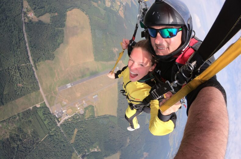 A selfie of a woman and man doing a tandem skydive with the airfield way below them