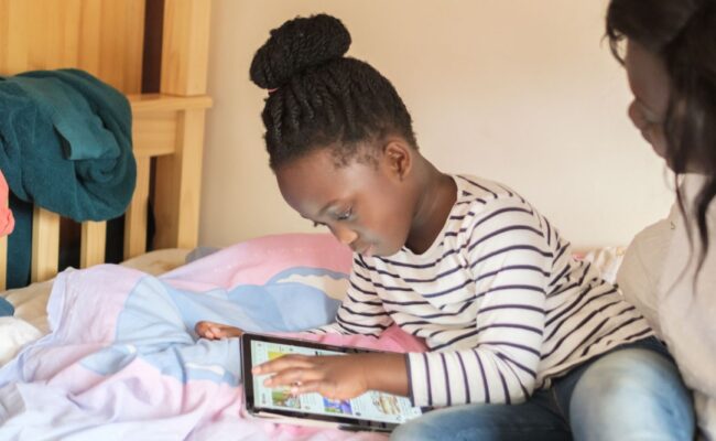 A mum and her daughter sit on the bed playing on the ipad together