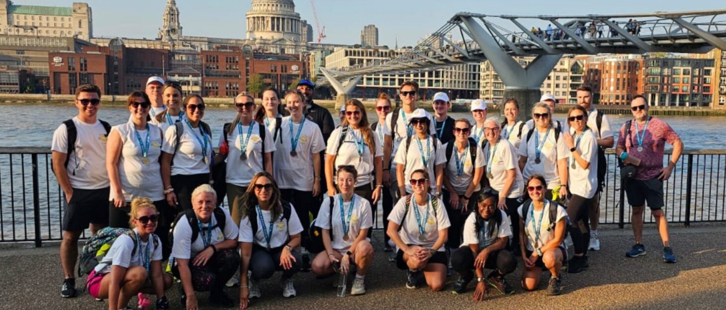 Large group of walkers in hiking equipment stood by Millennium Bridge in London, with the River Thames and St Paul's Cathedral in the background.