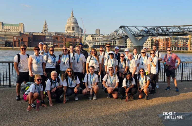 Large group of walkers in hiking equipment stood by Millennium Bridge in London, with the River Thames and St Paul's Cathedral in the background.