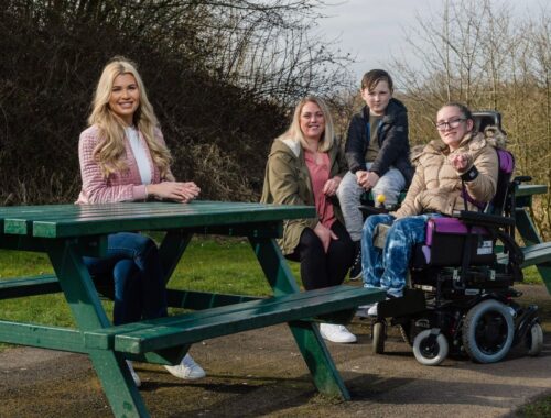 Woman sat on a park bench, with child in a wheel chair next to them and two other children sat down.