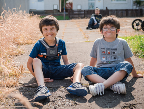 Two boys sit on the floor outdoors, smiling at the camera