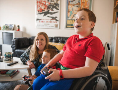 A young boy in a wheelchair plays on a games console with his younger brother, whilst mum watches on smiling.