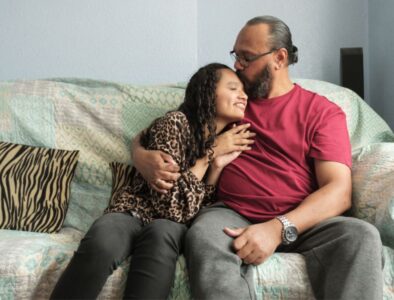 A blind girl and her father sat on a sofa, he kisses the top of her head