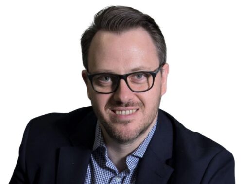 A man with dark hair and framed glasses wearing a navy blazer