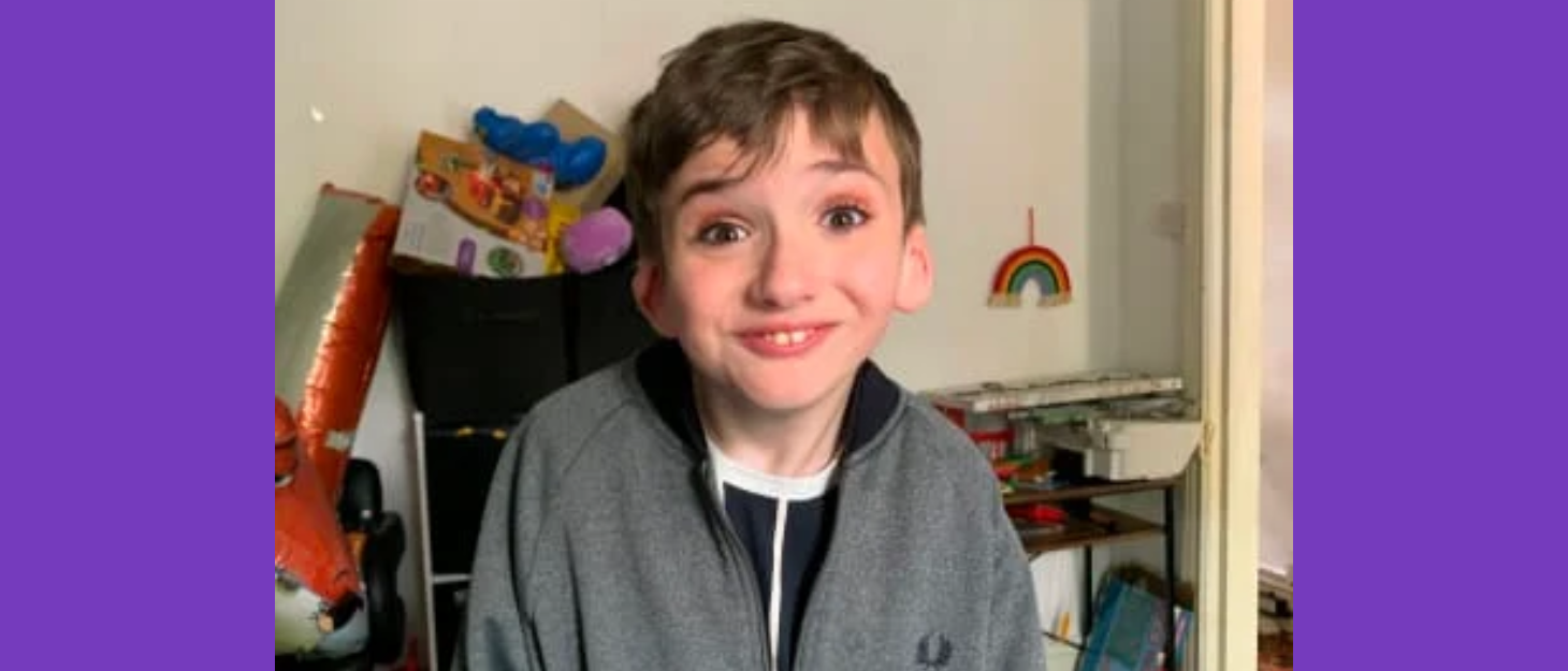 A young boy with eye shadow on standing in a bedroom smiling, wearing zip up hoodie