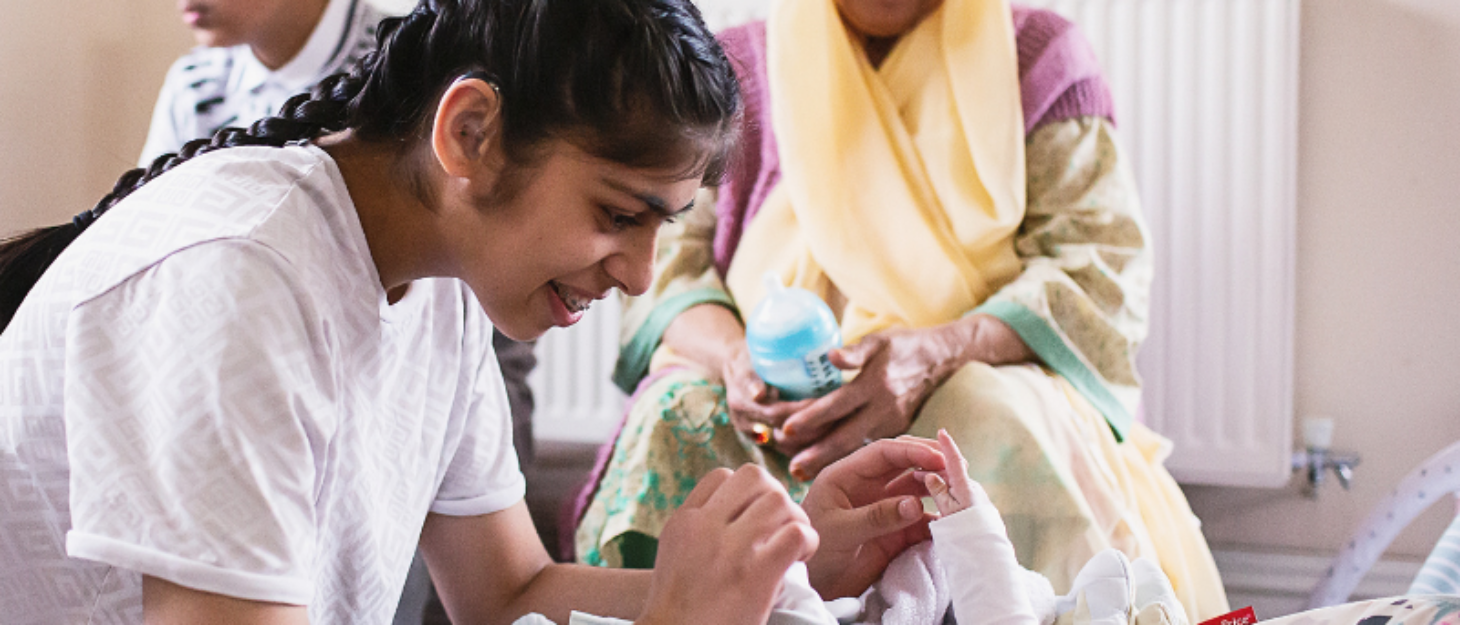 a girl playing with her baby sister while their brother and mum look over them