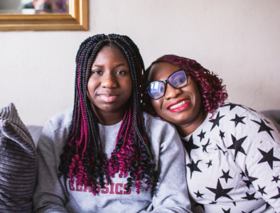 A teenage girl with long hair, and her mum seated next to here on a sofa smiling