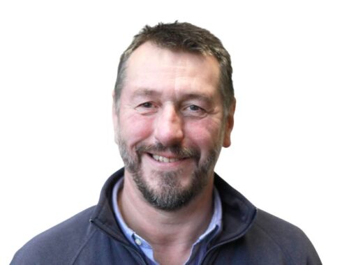 A man with a shirt and navy pull over, dark hair and short beard