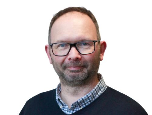 A man with a check shirt and navy jumper with dark framed glasses