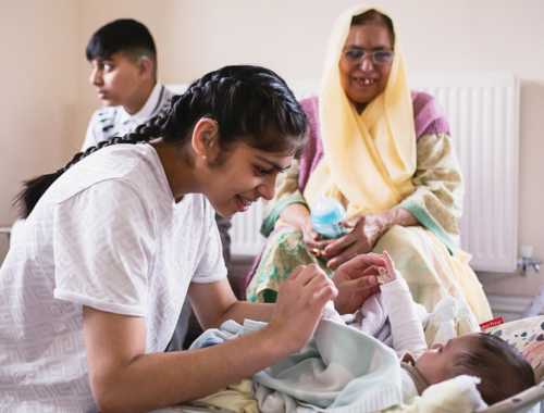 An older sister, with dark plaits and wearing a white t-shirt is playing with her younger sister in a baby chair. Their grant mother is in the background, smiling at them, next to their brother who is looking through the window