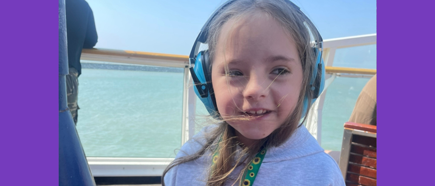 Young girl stands on boat wearing sunflower lanyard and ear defenders