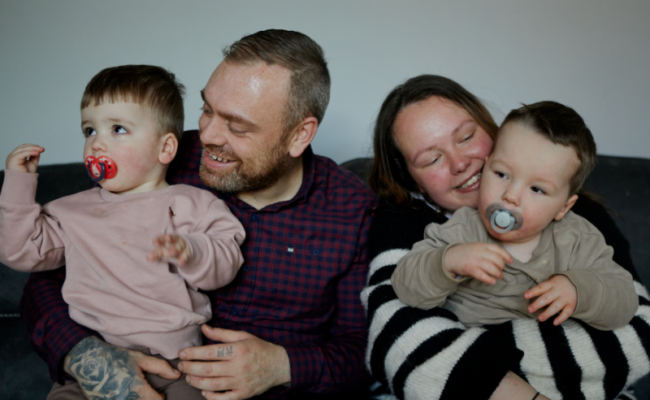 A family sitting on the sofa cuddling together. Dad holding the youngest son and mum holding the two year old son