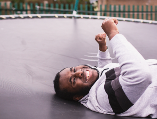 A teenage boy lies on his back on a trampoline. He is smiling and his hands are raised in the air.